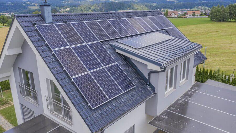 Modern house with a slate roof covered in solar panels, surrounded by a green field and distant trees under a clear sky, suggesting sustainable living.
